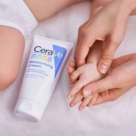 A tube of CeraVe Baby Moisturizing Cream lies on a soft white surface next to hands gently applying white cream to a baby's hand.